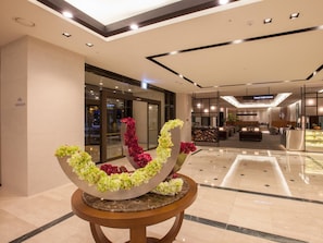 A lobby with a modern design, featuring a central floral arrangement on a round table, a seating area, and a visible entrance to a lounge area.
