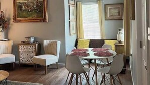 Un comedor con una mesa redonda, cuatro sillas y una ventana con cortinas. Hay un cuadro en la pared y una mesita auxiliar con un adorno.