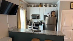 Cocina compacta con armarios blancos, un refrigerador de acero inoxidable, un microondas y una cafetera. Hay dos taburetes de madera en la barra.