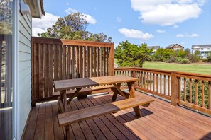 Una terraza de madera con mesas de picnic y barandilla, con vista a un área verde con casas al fondo.