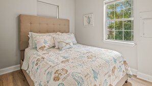 A bedroom with a large bed, a ceiling fan, a window with a view of trees, and a framed picture on the wall.