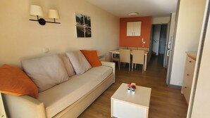 A living room with a sofa, a small table with flowers, a dining area with chairs, and a kitchen area with cabinets. - Comfortable nest with pool and sea view (Saint-Raphaël)