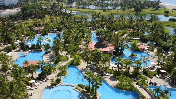 Ein Resort mit mehreren Schwimmbecken, umgeben von Palmen und Blick auf den Ozean.