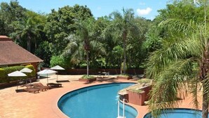 A swimming pool with a curved design, surrounded by lounge chairs and umbrellas, with palm trees and a building in the background. - Marcopolo Inn Iguazu (Puerto Iguazú)