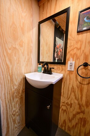 A bathroom with a wooden wall, a white sink, a black faucet, a mirror, and a framed picture. - Glamp in Heated Yurt in Big Bend @ Casa de Marca (Alpine)