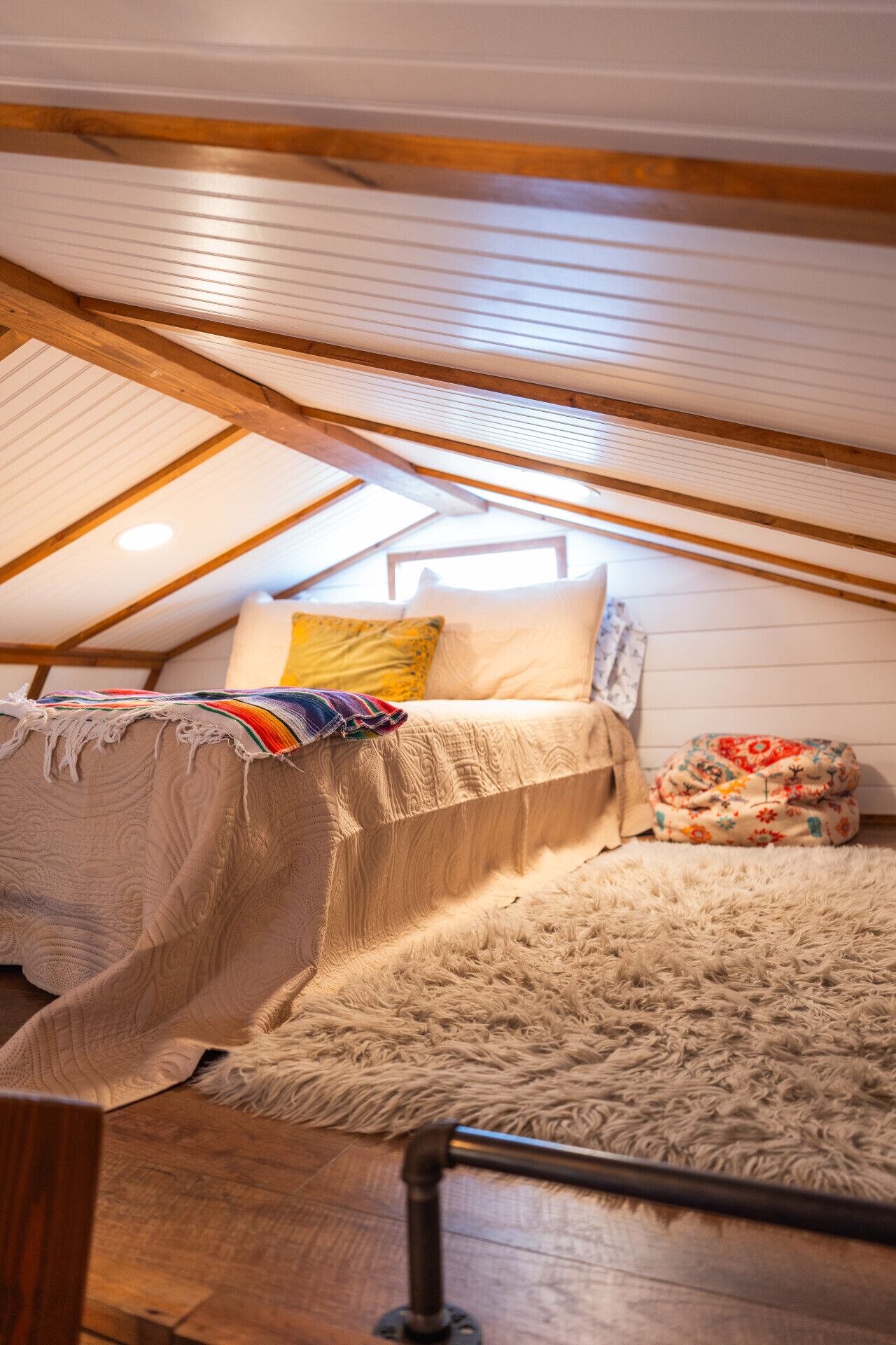 Un dormitorio acogedor en el ático, con techo inclinado, vigas de madera, una cama con manta colorida y una alfombra mullida.