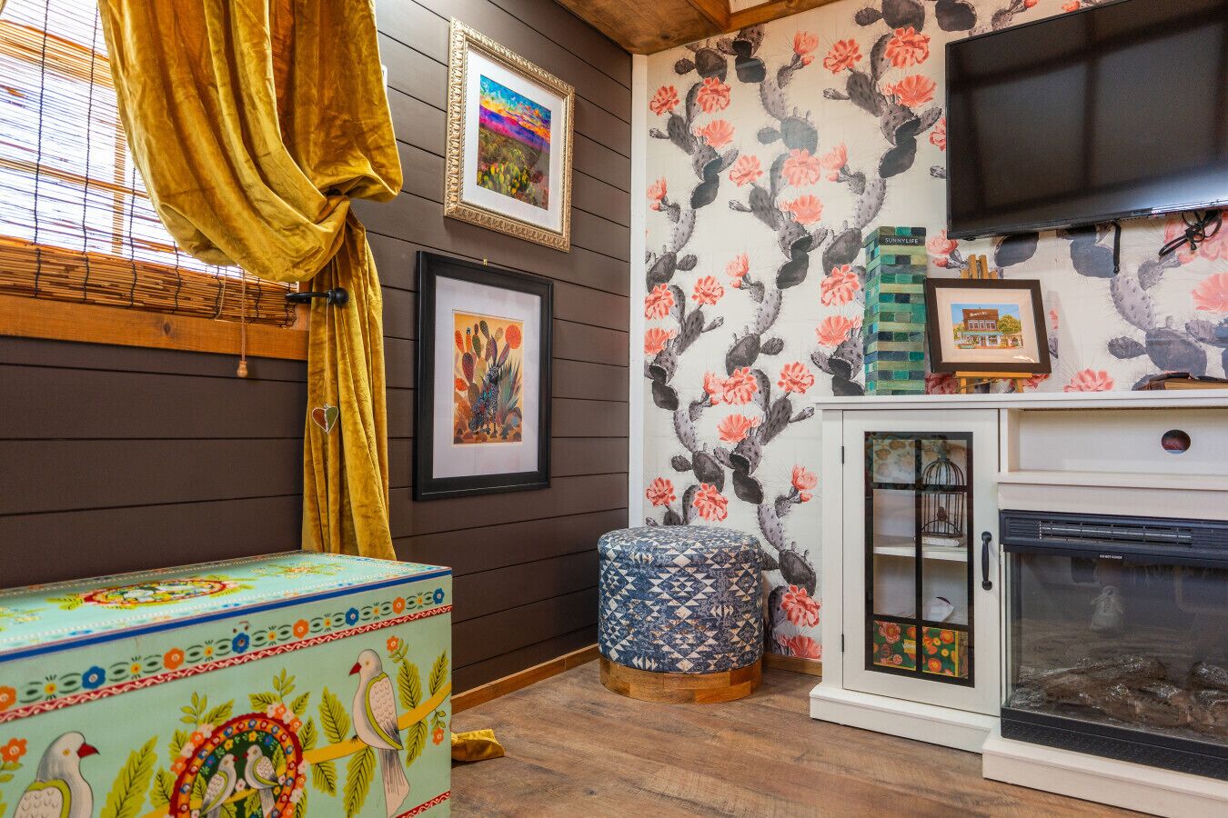 A cozy room with wooden walls, a fireplace, and a television. There are framed artworks and a colorful ottoman.