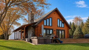 A log cabin with a stone chimney, a covered porch, and a deck with outdoor seating. - Juniper Presents: 15-Acre Retreat - Pool, Hot Tub, Pond, EV Charger (Rolling Prairie)