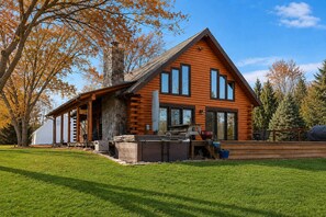 A log cabin with a stone chimney, a covered porch, and a deck with outdoor seating. - Juniper Presents: 15-Acre Retreat - Pool, Hot Tub, Pond, EV Charger (Rolling Prairie)