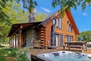 A log cabin with a hot tub, wooden deck, and stone fireplace. - Juniper Presents Scout's Haven: 15-Acre Retreat, Pool, Hot Tub, Pond & EV (Rolling Prairie)