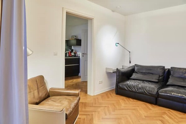 A living room with a sofa, a chair, and a lamp. There is a door leading to a kitchen area.