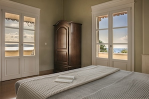 A bedroom with a large bed, a wooden wardrobe, and two windows with a view of the sea. - Villa Comi (Kefalonia)