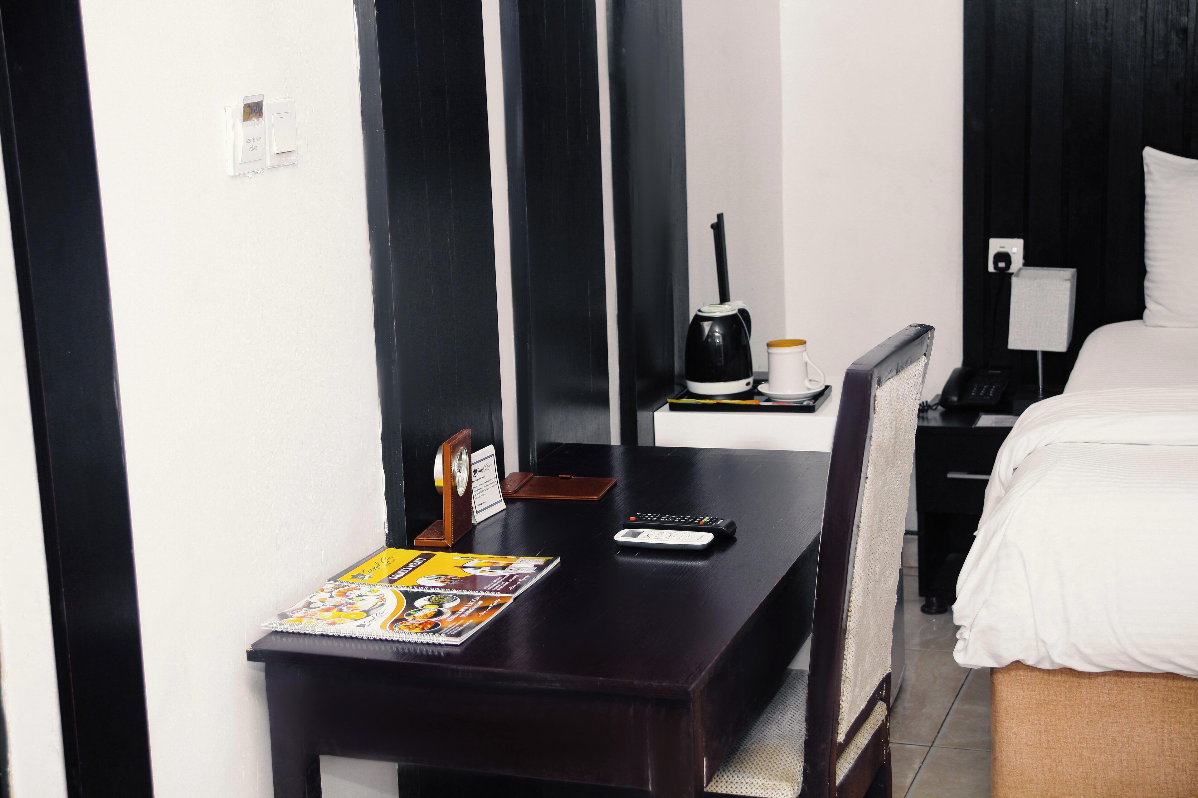 A hotel room with a dark wooden desk, a bed with white bedding, a nightstand with a lamp, and a coffee maker.