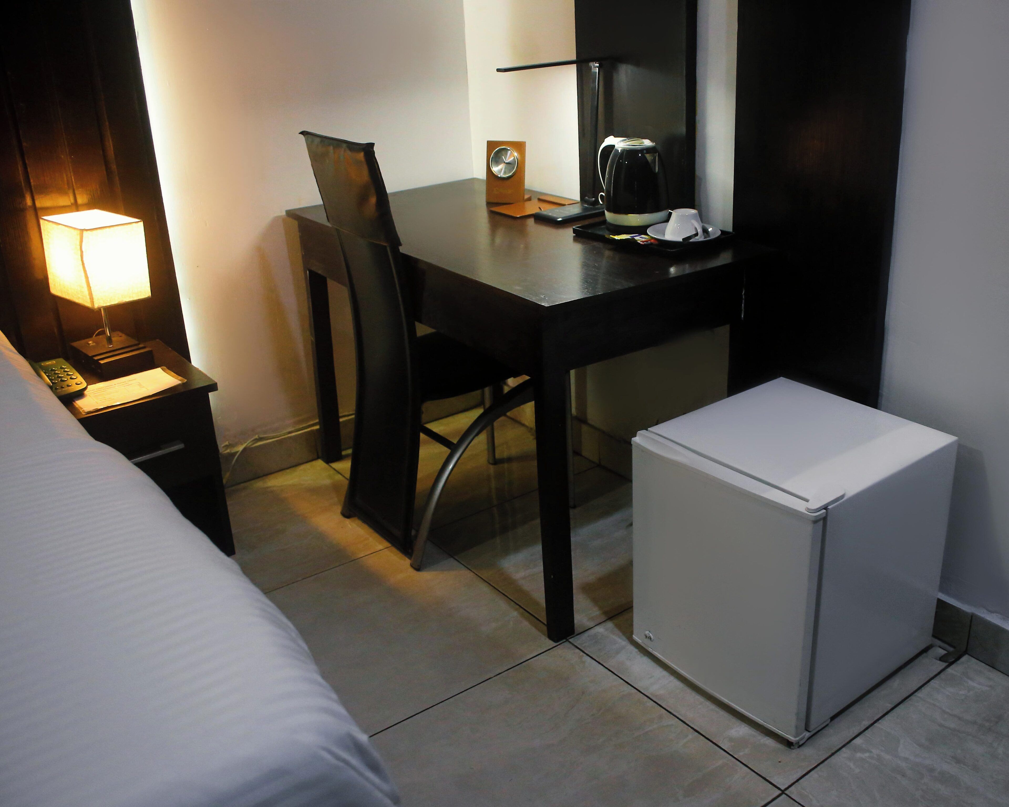 A hotel room with a bed, a desk with a lamp, a coffee maker, and a refrigerator.