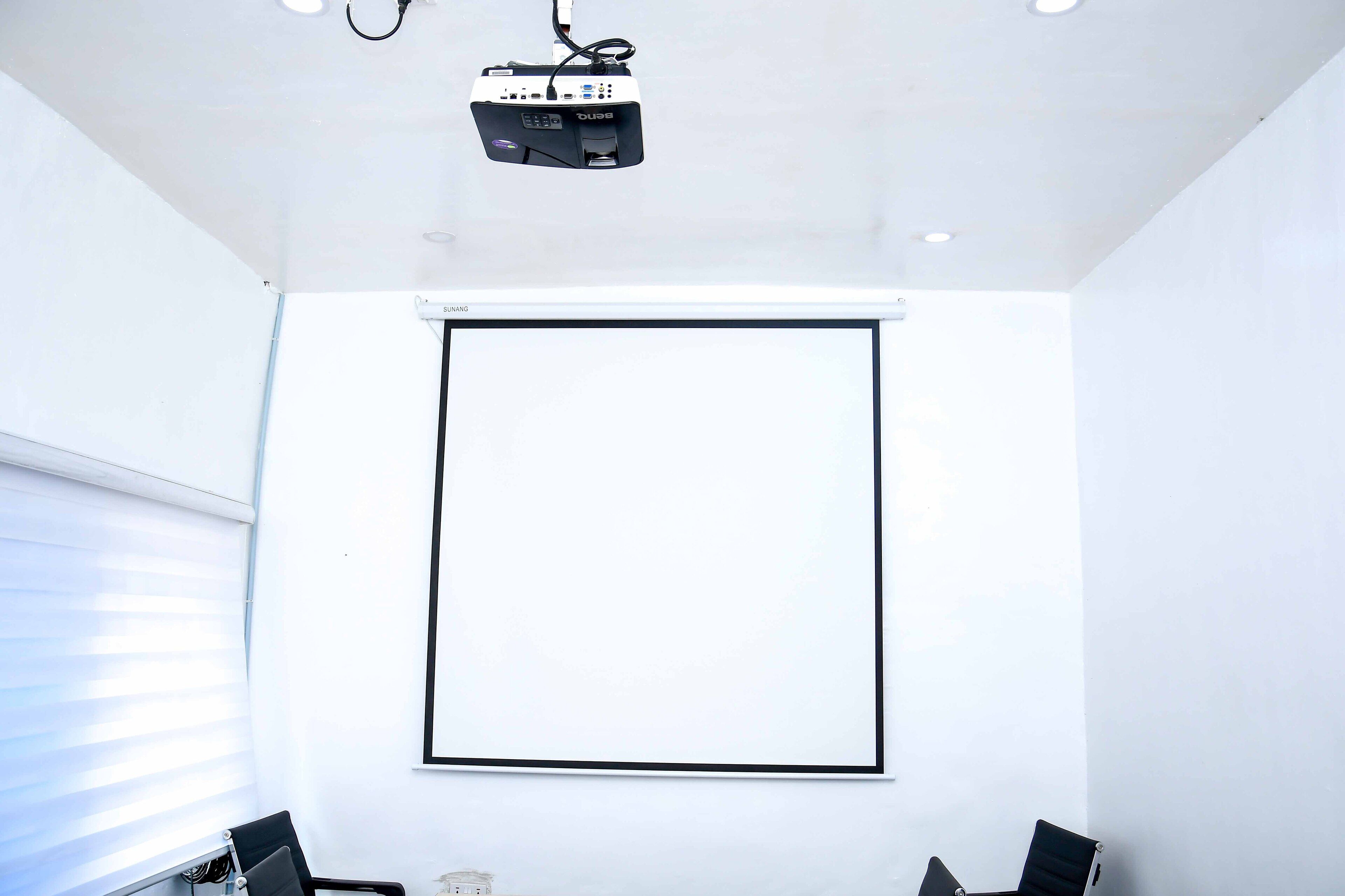Un salón de conferencias con un proyector, una pantalla en blanco y dos sillas.