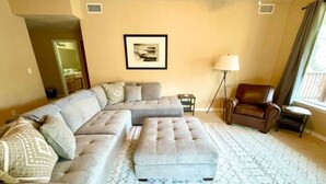 Una sala de estar con un sofá modular, un reposapiés, una lámpara de pie y un cuadro en la pared.