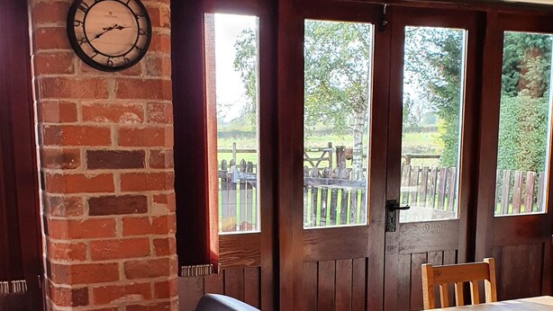 A cozy room with a leather sofa, a wooden dining table, and a brick wall with a clock. - Hayloft Barn - Lodge Barns , Country Get Away, Morton near Southwell (Nottinghamshire)