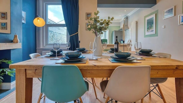 A dining area with a wooden table set for four, blue chairs, a vase with green plants, and a window with blue curtains. - 2-Bed Southsea House with Garden, Fireplace and Free Parking (Portsmouth)