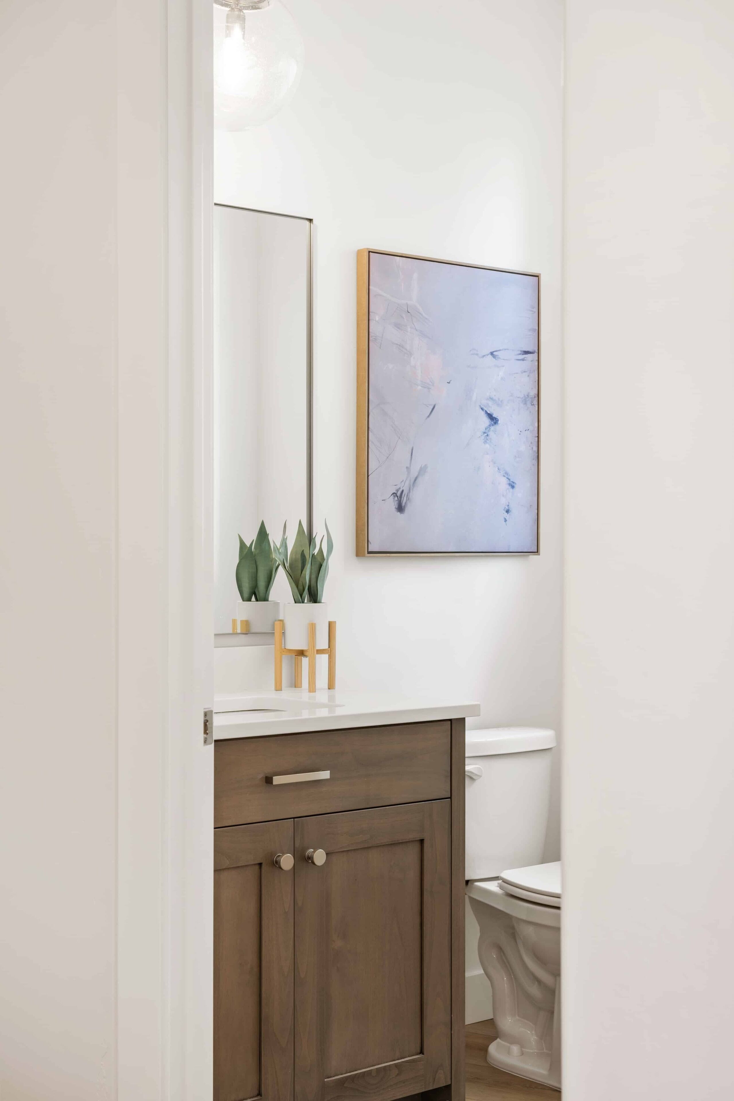 A bathroom with a white vanity, a wooden cabinet, a white toilet, and a framed abstract painting on the wall.