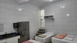 Eine kompakte Küche mit Kühlschrank, Spüle und Esstisch. Ein kleines Bett mit Kissen und ein Regal darüber.
