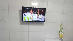 Ein Zimmer mit Fernseher, der einen Kampf der UFC zeigt, einer Mikrowelle, einem Wasserkocher und einem Bett mit blumigem Überwurf.