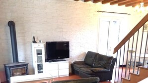 A living room with a black leather sofa, a television on a white cabinet, a black wood-burning stove, and a staircase with wooden beams. - Detached house in LE PARC DU MORVAN in Chalaux for 4 people. (Chalaux)