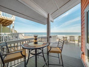 A covered outdoor area with a round table and chairs, overlooking a beach. - Ocean Dunes 1903 | Oceanfront, Spacious Layout, Views + Resort Amenities (Kure Beach)