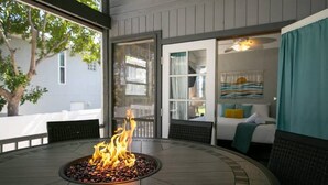 A round table with a fire pit in the center, a wicker chair, and a view of a bedroom with a bed and curtains.