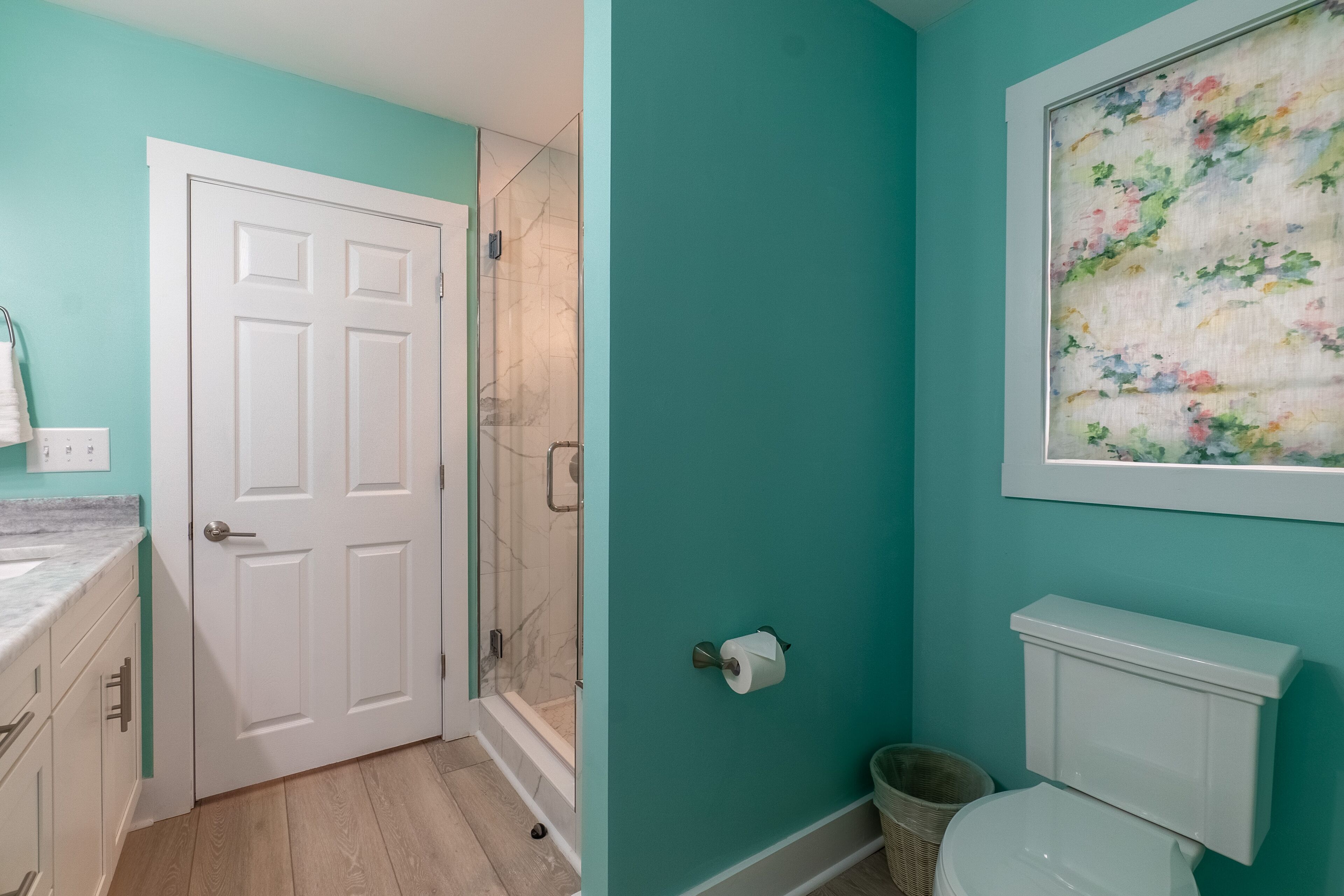 A bathroom with teal walls, a white toilet, a shower with glass doors, and a marble countertop.