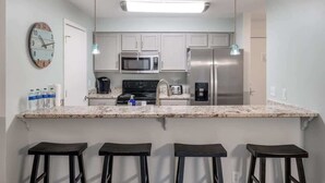 A modern kitchen with white cabinets, stainless steel appliances, a granite countertop, and three bar stools. - Relax & Enjoy Life! Updated Wild Dunes Condo (Isle of Palms)