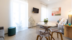 A modern living room with a glass dining table, a sofa, a TV mounted on the wall, and a large window with sheer curtains.