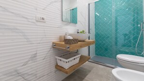 A modern bathroom with a white marble wall, a wooden shelf with toiletries, a white sink, and a glass shower enclosure.