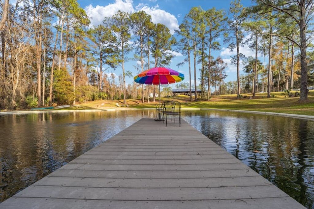 A wooden dock extending into a calm body of water, surrounded by tall trees and a colorful umbrella.