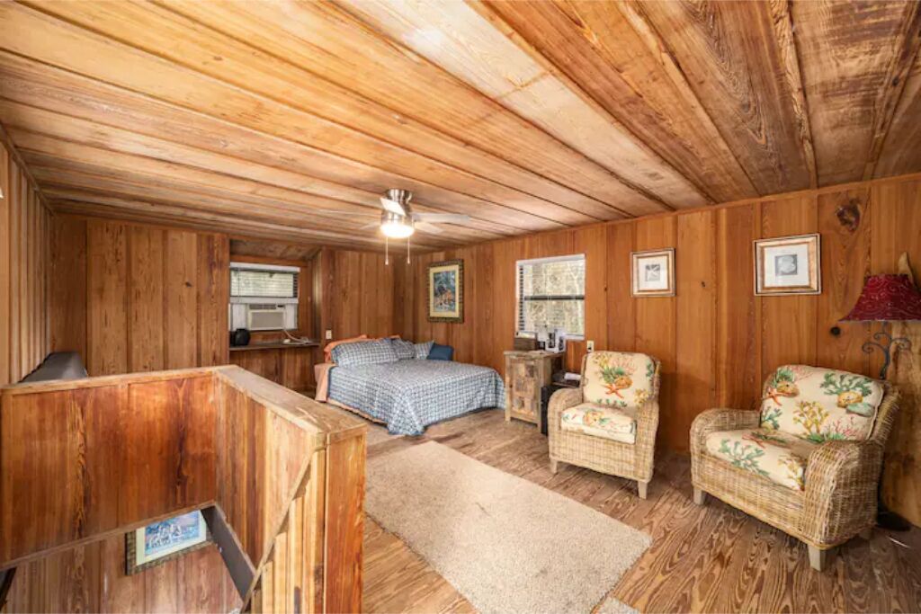 Une pièce chaleureuse aménagée dans un chalet, avec des murs en bois, un lit recouvert d’une courtepointe à carreaux bleus, deux fauteuils en osier, une petite table et un ventilateur de plafond.