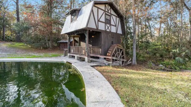 A rustic cabin with a balcony, a wooden deck, and a pond in the foreground. - Wilderness Getaway at The Grist Mill (Fort McCoy)