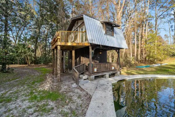 A rustic cabin with a wooden deck and a metal roof, surrounded by trees and a pond.