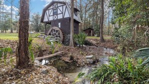 Une cabane rustique dotée d’une grande roue à eau, entourée d’arbres et bordée d’un petit ruisseau.
