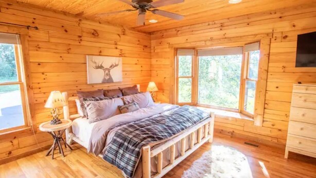 Un dormitorio de cabaña de madera con una cama, dos mesitas de noche, un ventilador de techo y una ventana con vistas al exterior.