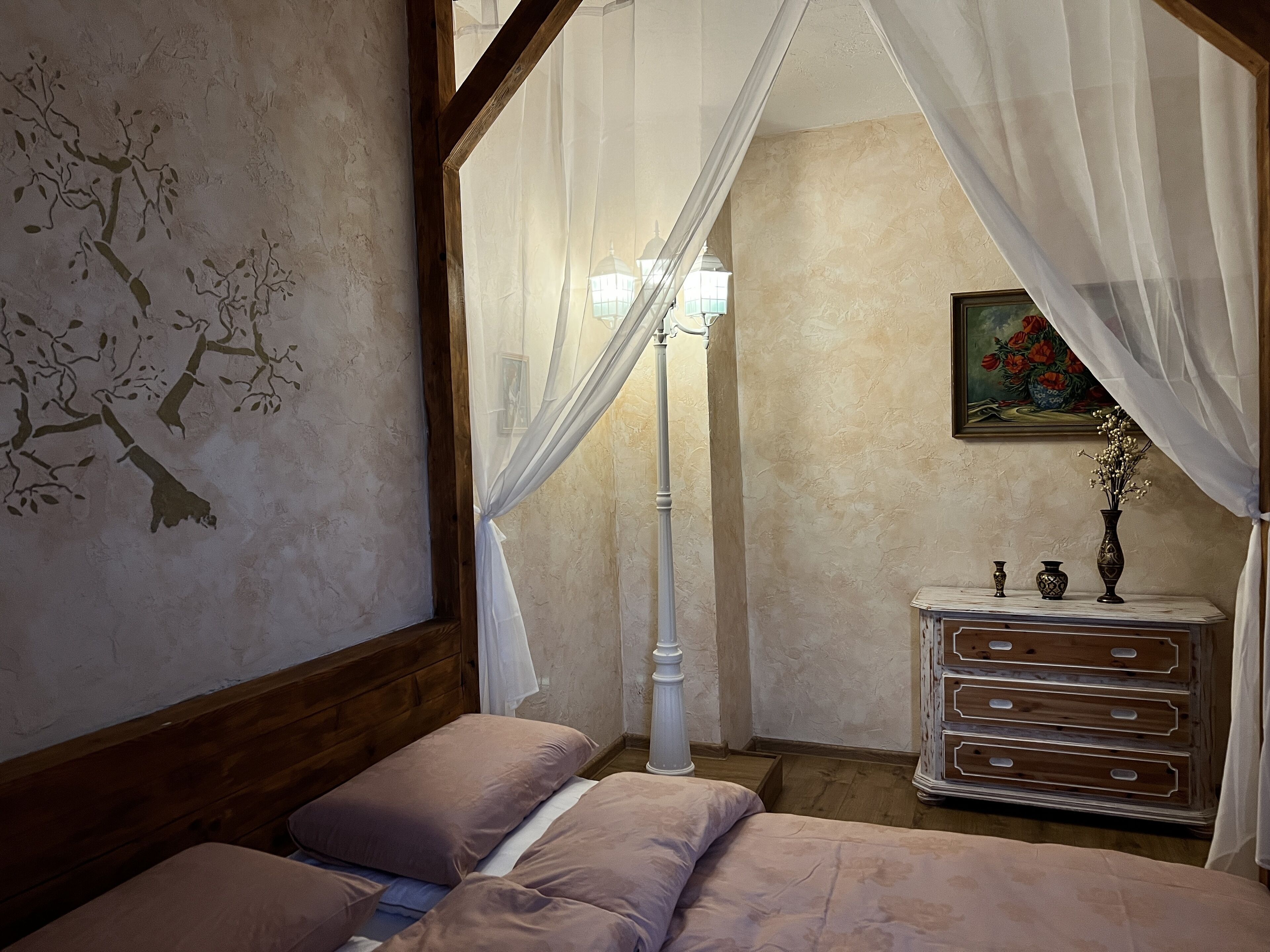 A bedroom with a canopy bed, a bedside table with decorative items, and a wall painting.