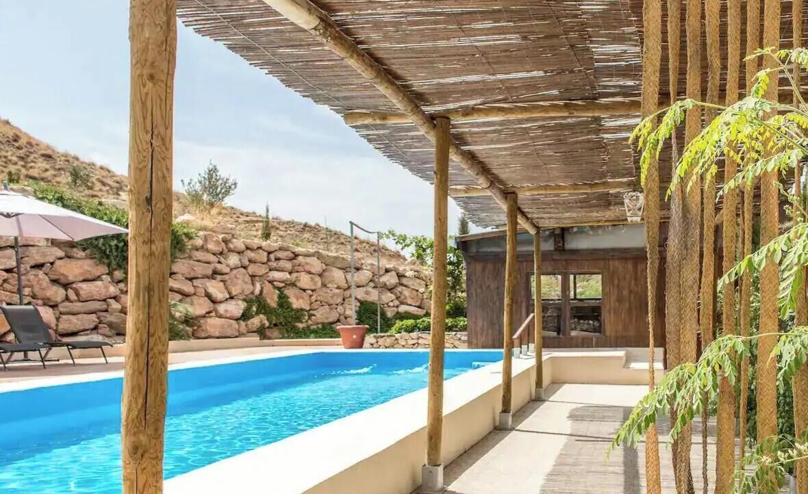 A pool area with a stone wall, wooden pergola, and a lounge chair.