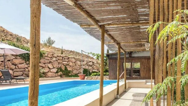 A pool area with a stone wall, wooden pergola, and a modern building in the background.