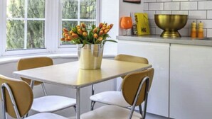 A kitchen with a dining table, chairs, a yellow pendant light, a vase with flowers, a white brick backsplash, and a window with blinds. - 70s Inspired, 3-Bed Home in Rye with river views (Rye)