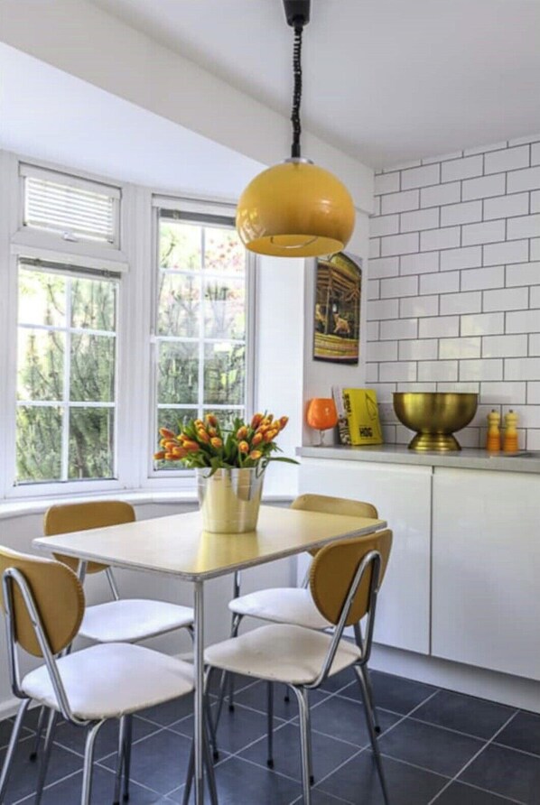 Une cuisine avec une table à manger, des chaises, un luminaire suspendu jaune, un vase avec des fleurs, un mur de briques blanches derrière l’évier et une fenêtre avec des stores.