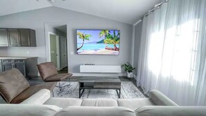 A modern living room with a large flat-screen TV, a sofa, a coffee table, and a kitchen area in the background.