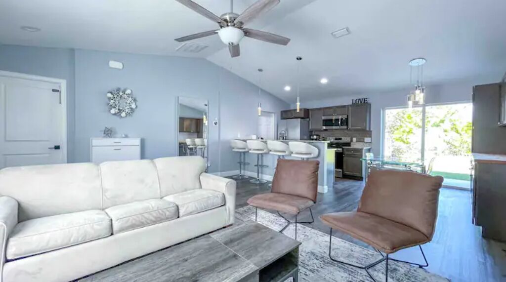 A modern living room with a white sofa, two brown chairs, a coffee table, and a ceiling fan. A kitchen with bar stools is visible in the background.