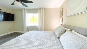 A bedroom with a large bed, a ceiling fan, a TV mounted on the wall, and a window with curtains.