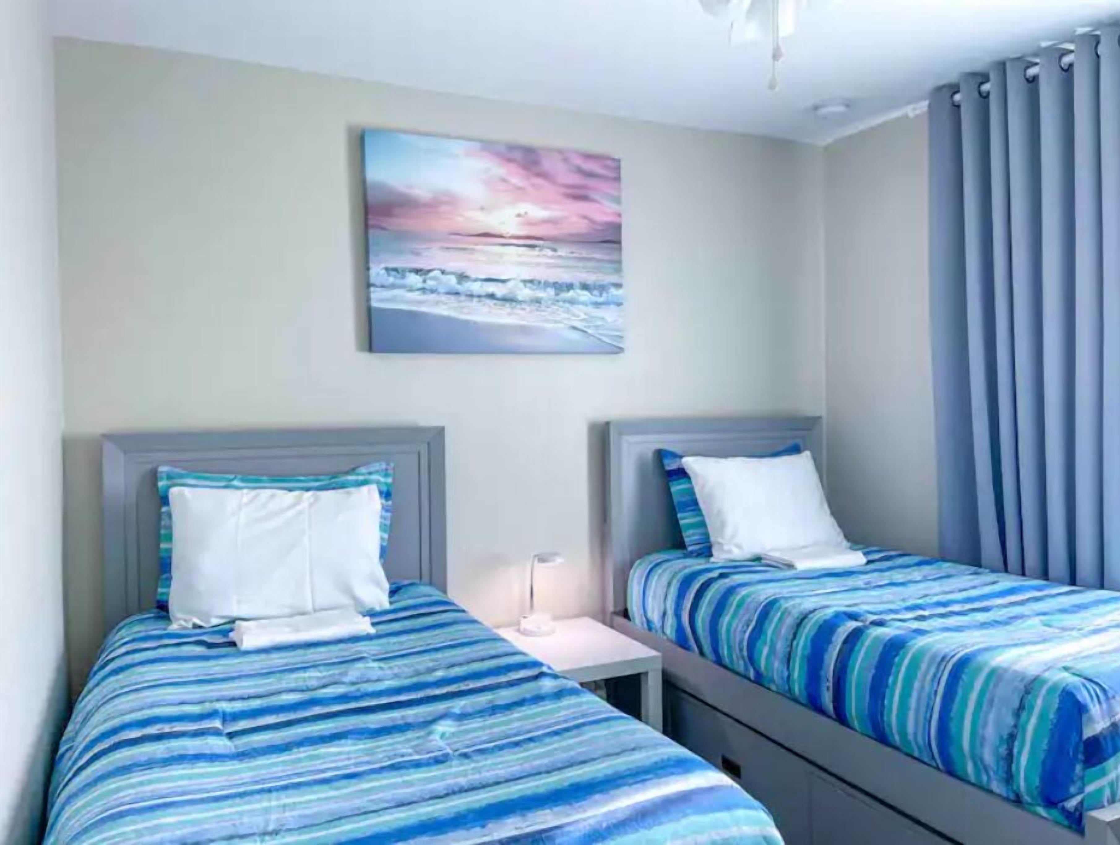 Een hotelkamer met twee bedden, elk voorzien van een blauw-wit gestreept laken, een nachtkastje, een bijzettafeltje, en een schilderij van een zonsondergang boven de oceaan aan de muur.