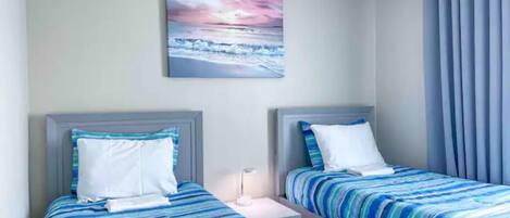 Een hotelkamer met twee bedden, elk voorzien van een blauw-wit gestreept laken, een nachtkastje, een bijzettafeltje, en een schilderij van een zonsondergang boven de oceaan aan de muur.