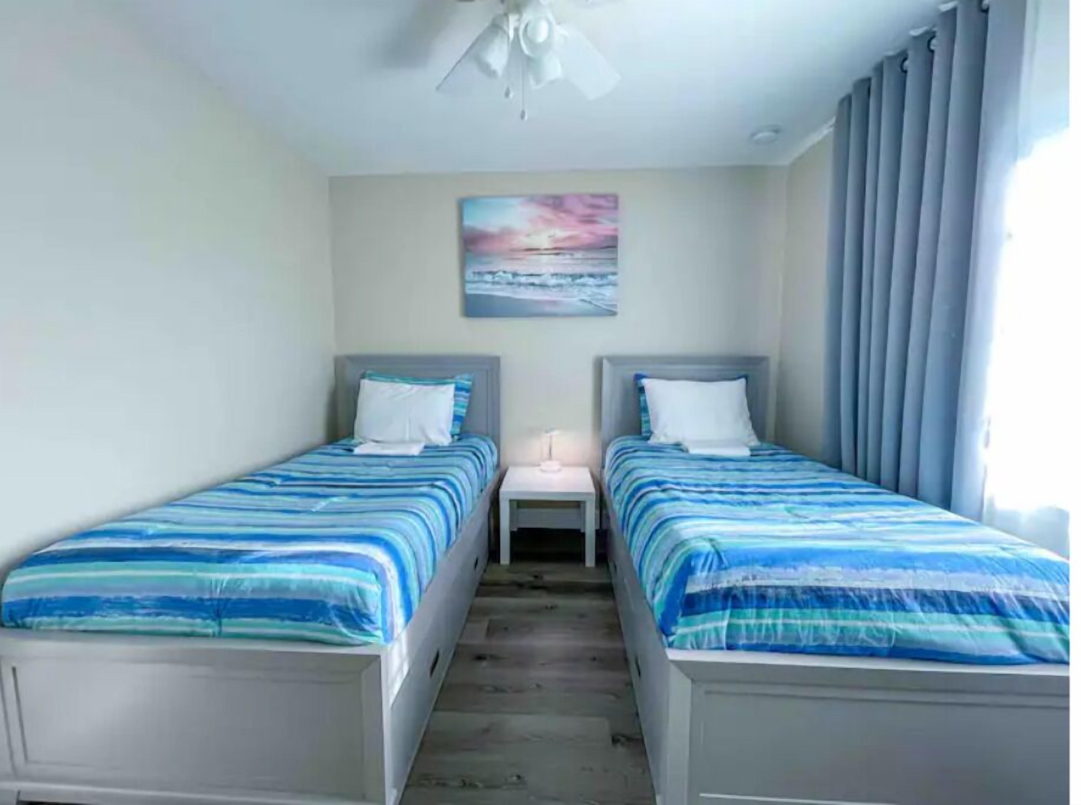 Quarto com duas camas, uma mesinha de cabeceira, um ventilador de teto e um quadro na parede.