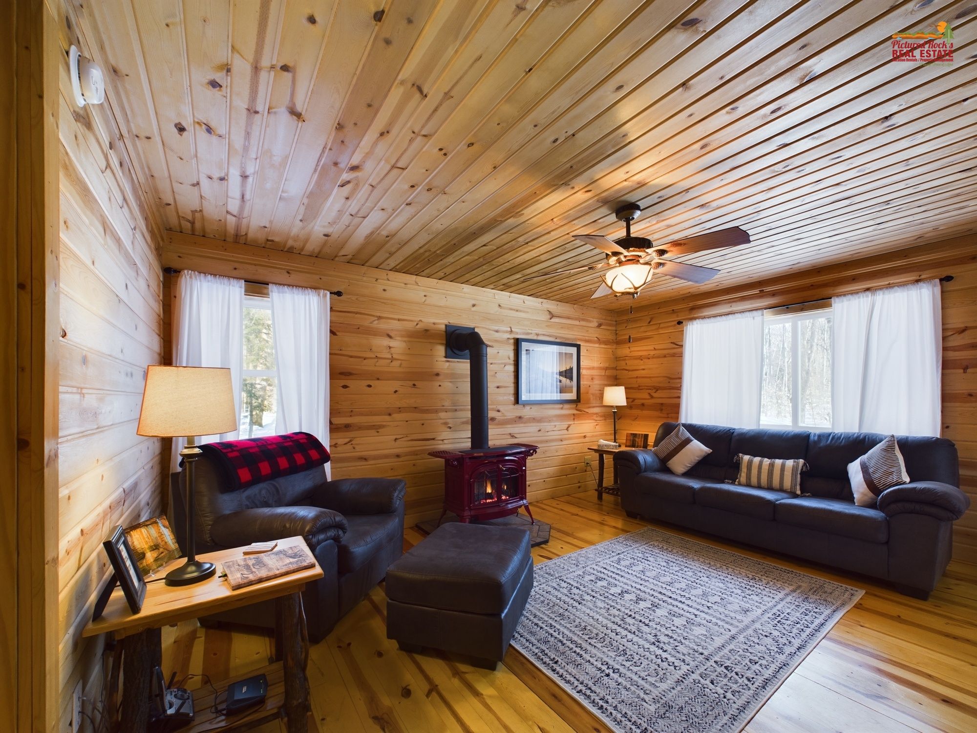 A cozy cabin interior with wooden walls, a ceiling fan, a fireplace, two sofas, a coffee table, and a rug.